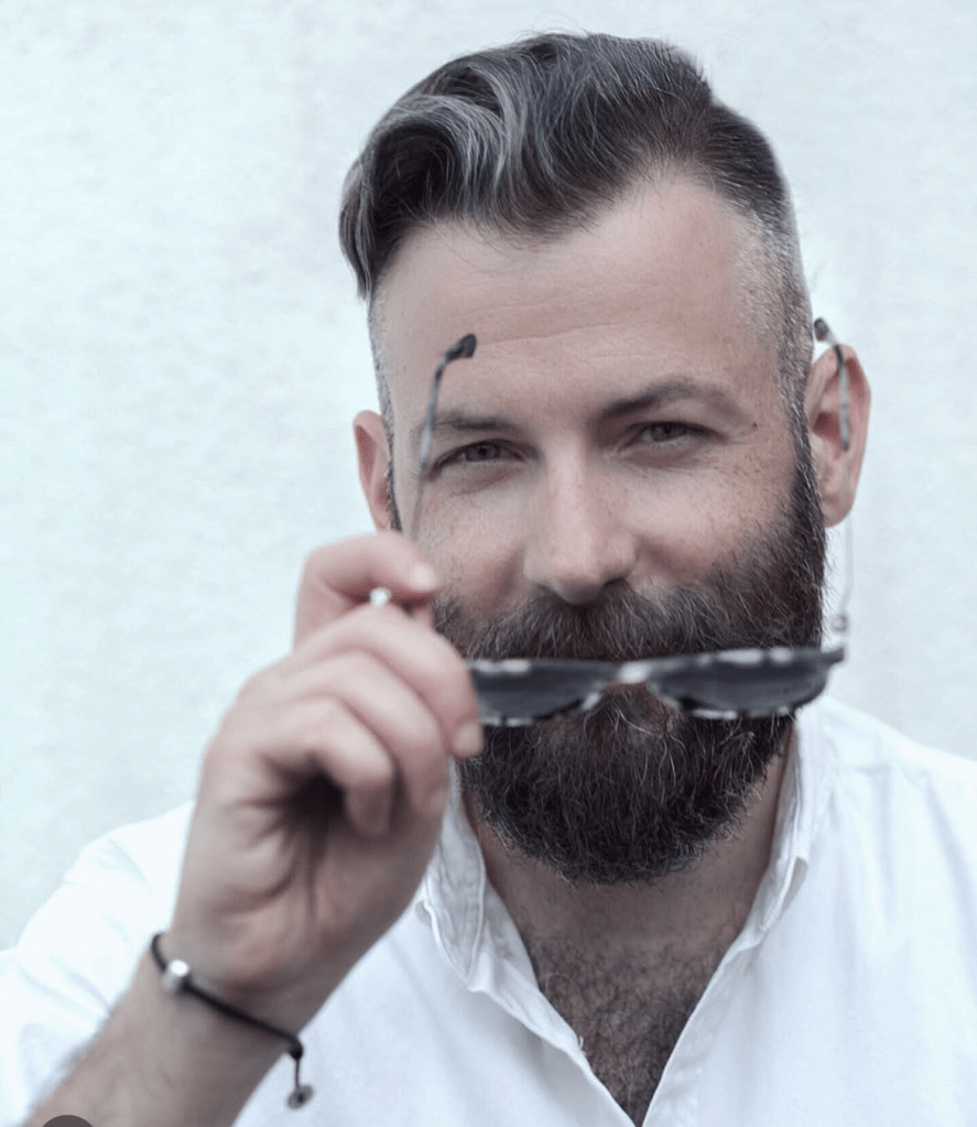 Portrait of Deniz Kosan with full beard and slicked-back hair, holding sunglasses